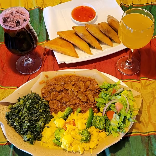 Ethiopian platter with injera, various stews, vegetables, samosas, and two drinks on a colorful tablecloth.